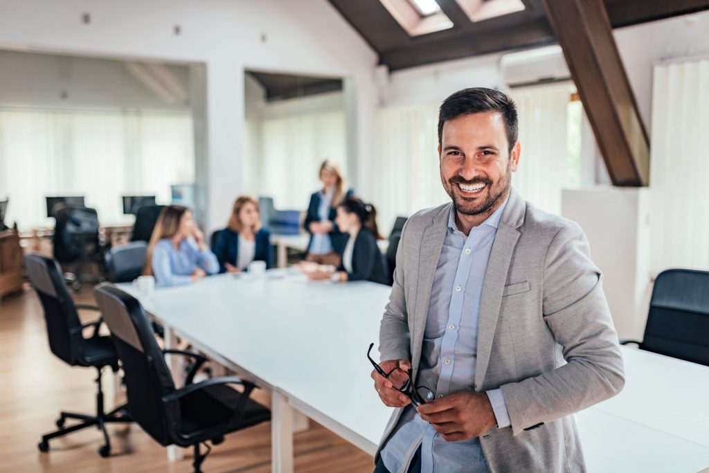 Business man holding his glasses, and smiling at the camera while standing infront of meeting table where business women are in discussion.