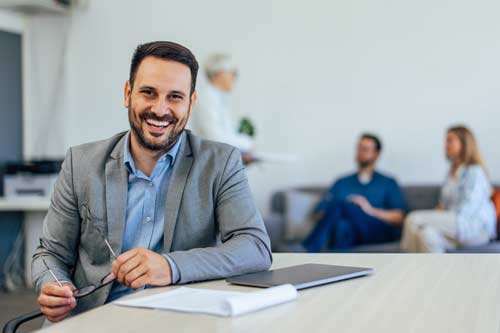 Business man holding his glasses, and smiling at the camera while sitting infront of meeting table where people are in discussion.